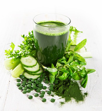 bicchiere di frullato verde clorella in mezzo a verdura verde e capsule verdi di clorella